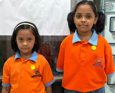 Two young girls in orange school uniforms with smiley face badges.