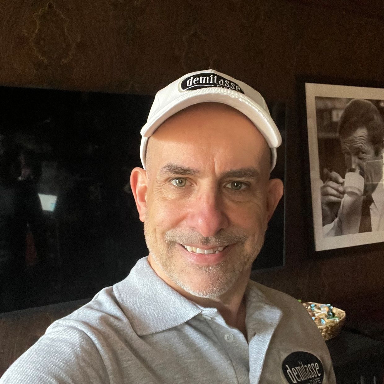 Man wearing a white cap and gray polo shirt with "demitasse" logo smiling indoors.