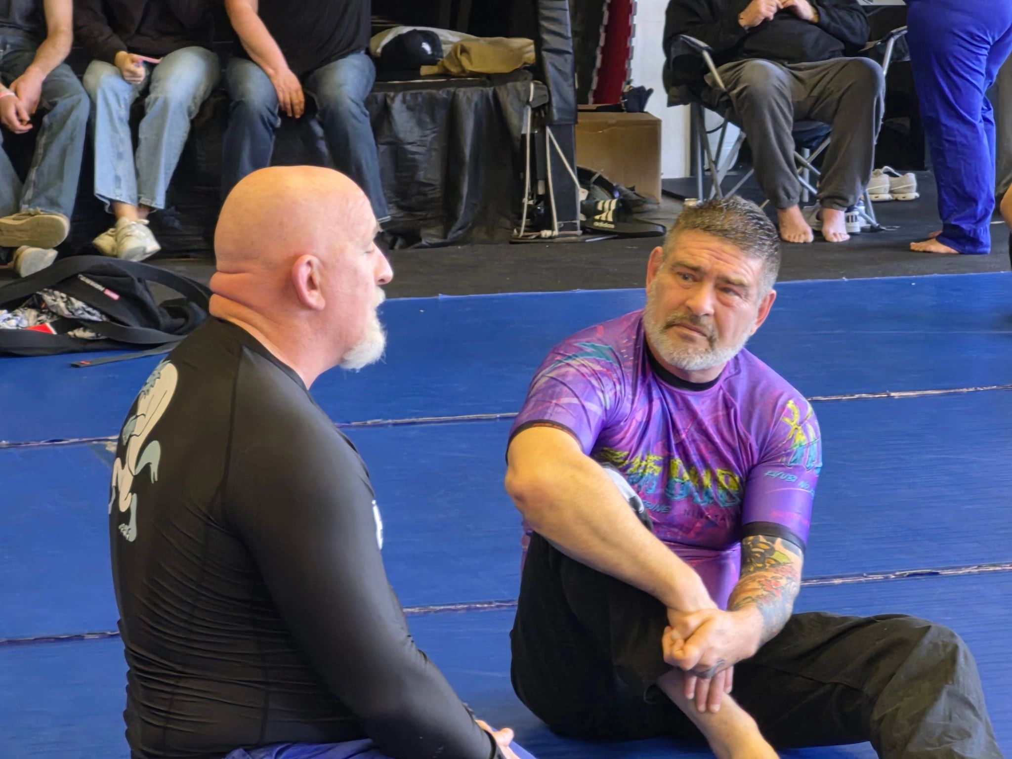 Two men in martial arts attire sitting on a mat, engaged in conversation.