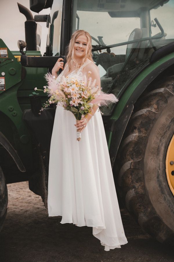 Bride in white dress holding a bouquet, standing next to a tractor.