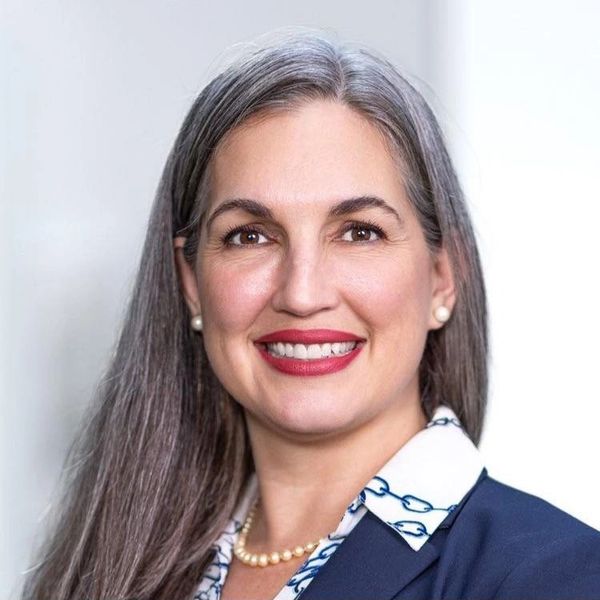Photo of a smiling woman, with long grey and black hair, red lipstick and blue suit looking unapolog