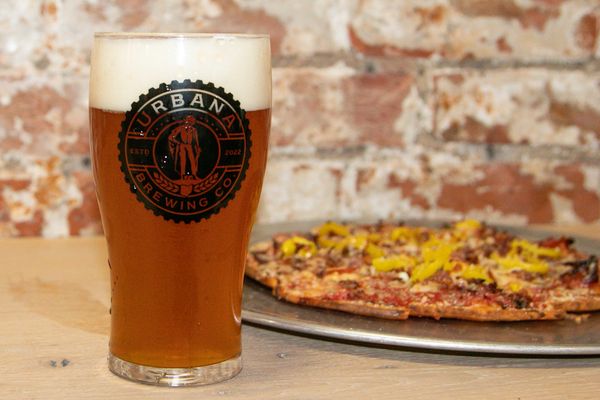 Pint of craft beer served on a wooden table, paired with a fresh pizza in the background.