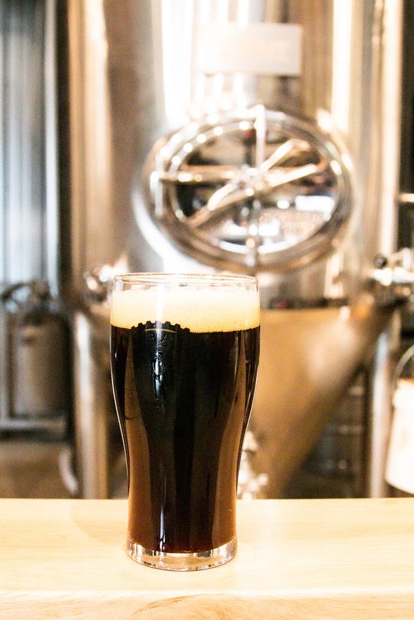 A pint of dark craft beer with a foamy head on a wooden bar.
