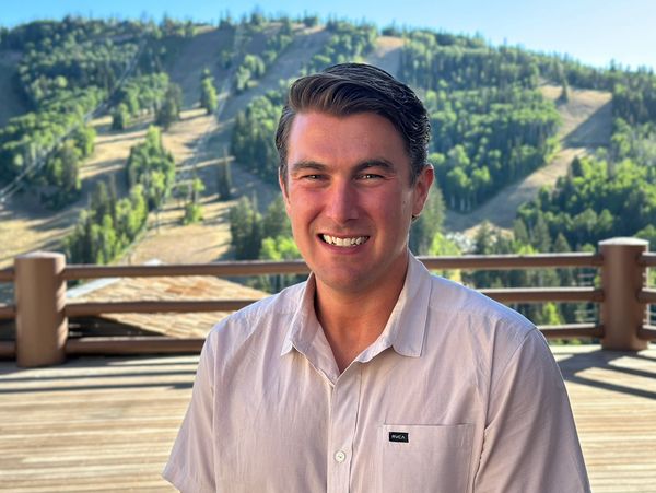Smiling man in a light shirt on a sunny balcony with forested hills behind.