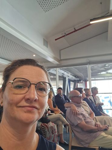 Woman taking selfie in a room with seated people in the background.