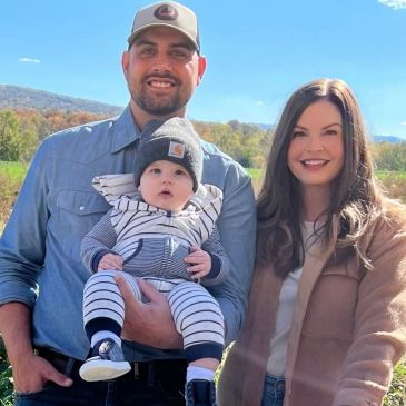 A happy family posing outdoors on a sunny day with a baby in striped outfit.