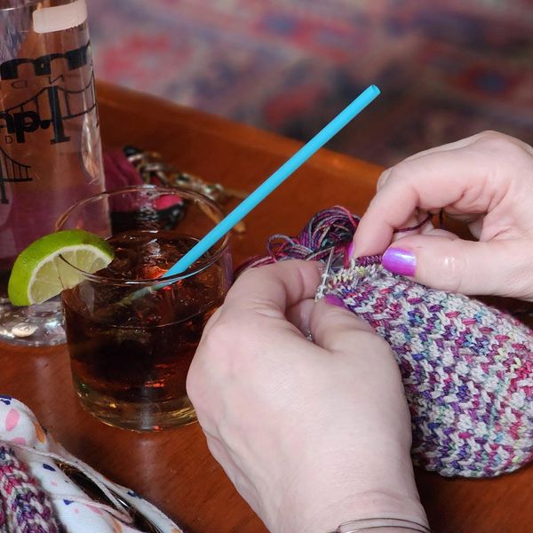 Hands knitting a colorful project beside a drink with a lime wedge.