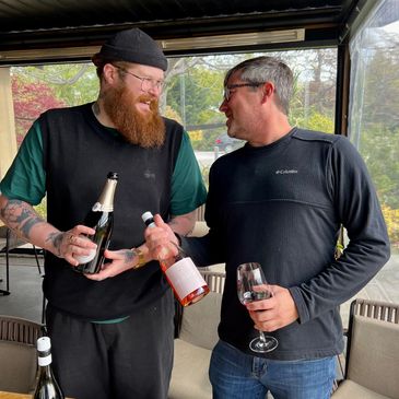 Two men sharing drinks and smiling indoors at a wooden table.