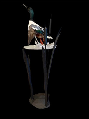 Artistic metal sculpture of a duck perched on a circular platform with a dark background.