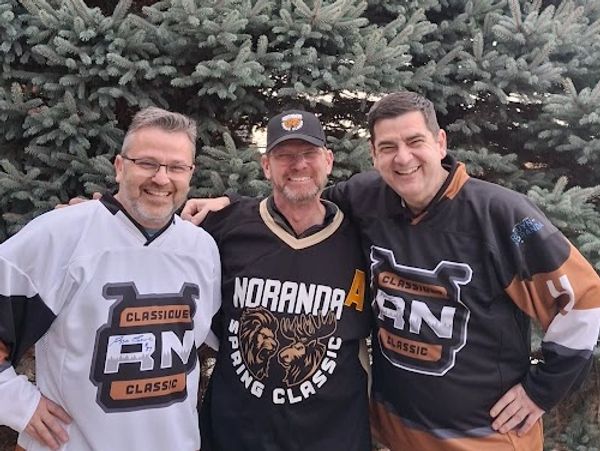 Three men in hockey jerseys smiling outdoors in front of pine trees.