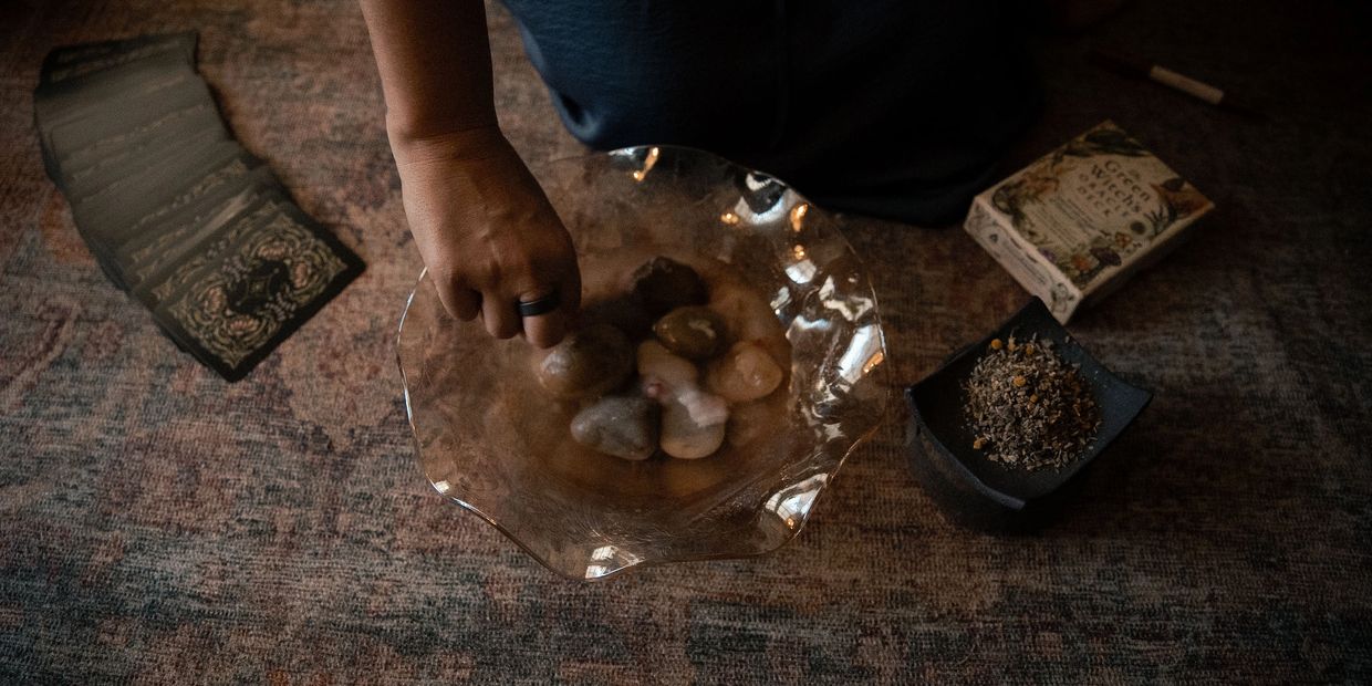 Person holding stones in a glass bowl with tarot cards and herbs nearby.