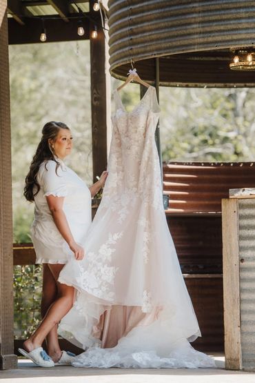 Bride in white robe admiring her hanging wedding dress outdoors.