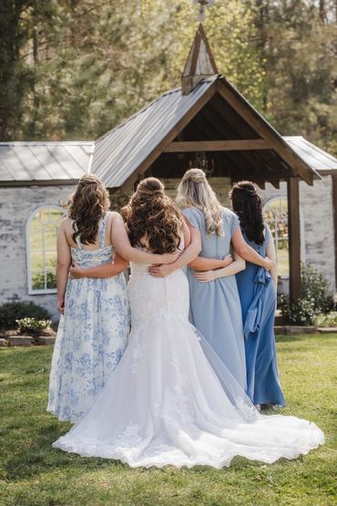Bride with bridesmaids embracing in front of a rustic chapel outdoors.