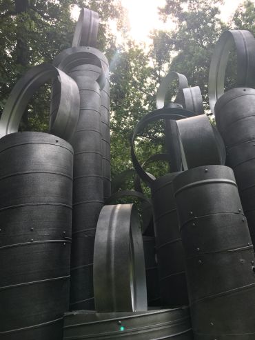 Tall, cylindrical metal sculptures with looped tops under a bright sky and surrounded by trees.
