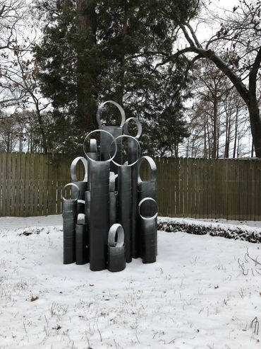 Black metal sculpture in snow-covered yard with trees and wooden fence.