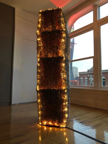 Tall, illuminated rectangular sculpture with warm lights in a bright room.