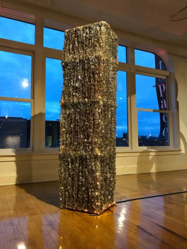 Tall sculpture made of metallic chains illuminated in a gallery room.