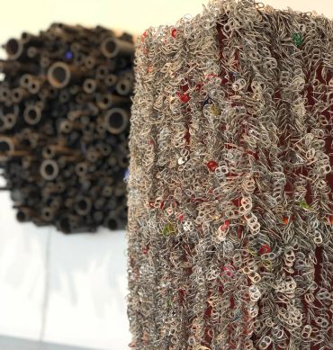 Close-up of intricate sculpture made from metal pull tabs with blurred circular art in background.