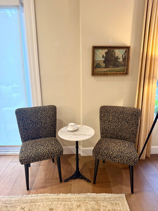 Two leopard print chairs flanking a marble top table with a cup, under a landscape painting.