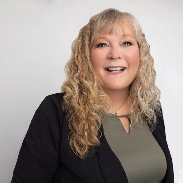Smiling woman with curly blonde hair in business attire against a plain background.