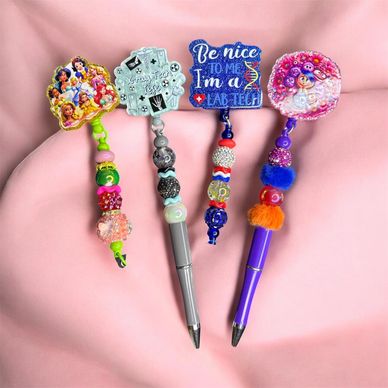 Colorful decorated pens with unique toppers and beads on a pink fabric background.