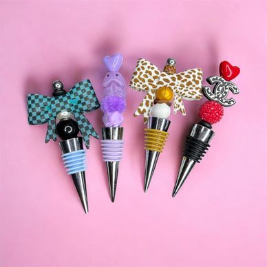 Four decorative wine bottle stoppers with colorful tops on a pink background.