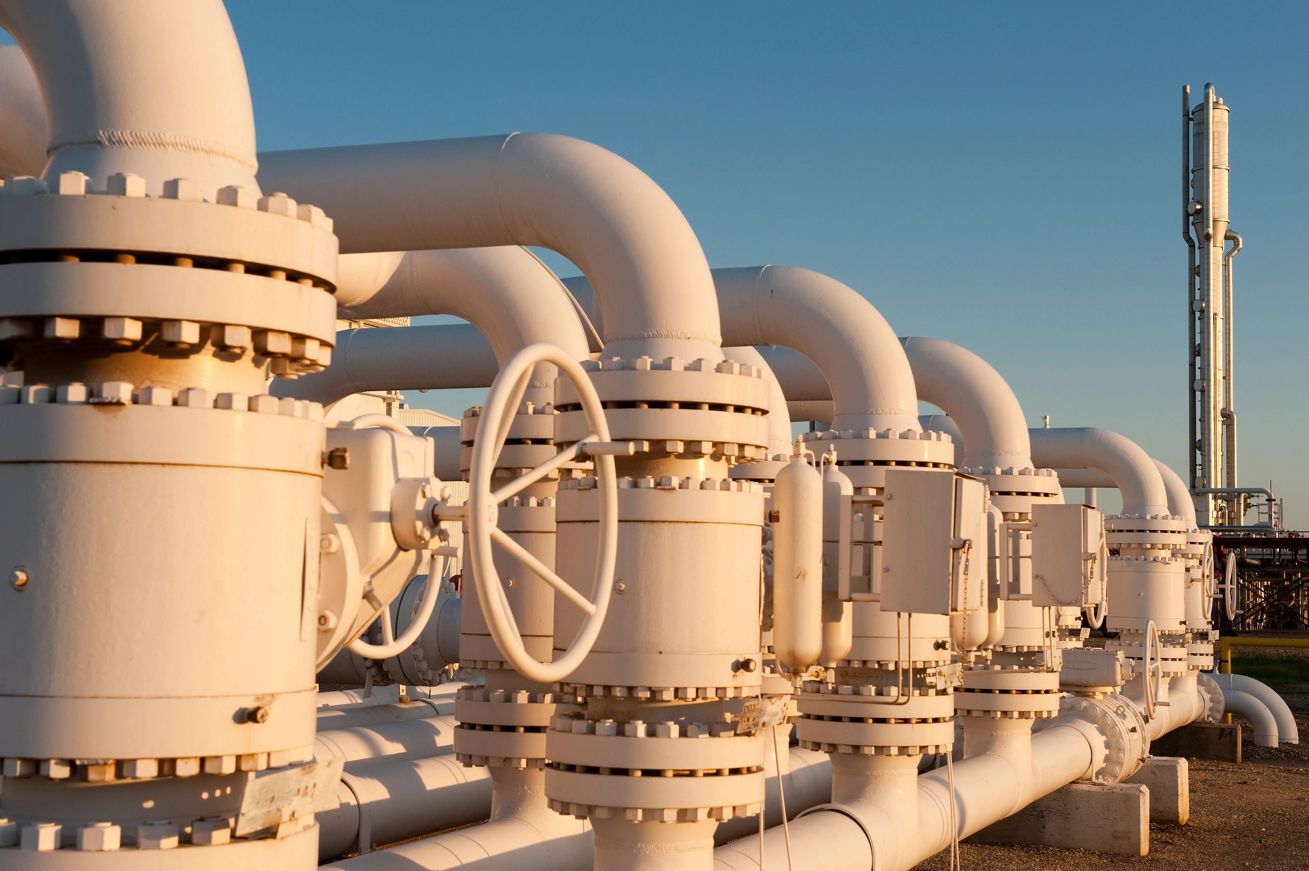 Large white industrial pipes and valves at a gas facility during sunset.