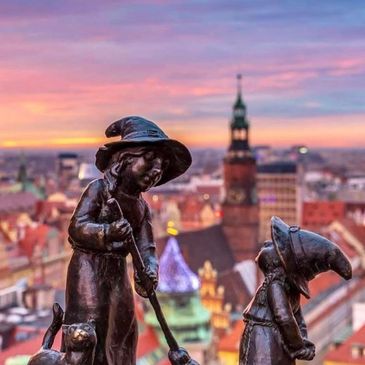 Bronze statues of two children wearing hats at sunset with cityscape.