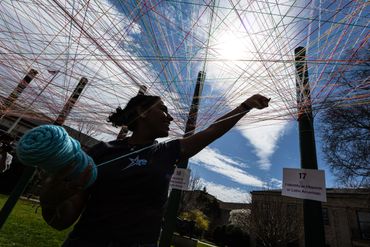 Person weaving colorful threads in an outdoor art installation under a bright sky.
