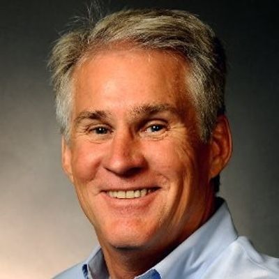 Smiling middle-aged man with gray hair and blue shirt against a dark background.