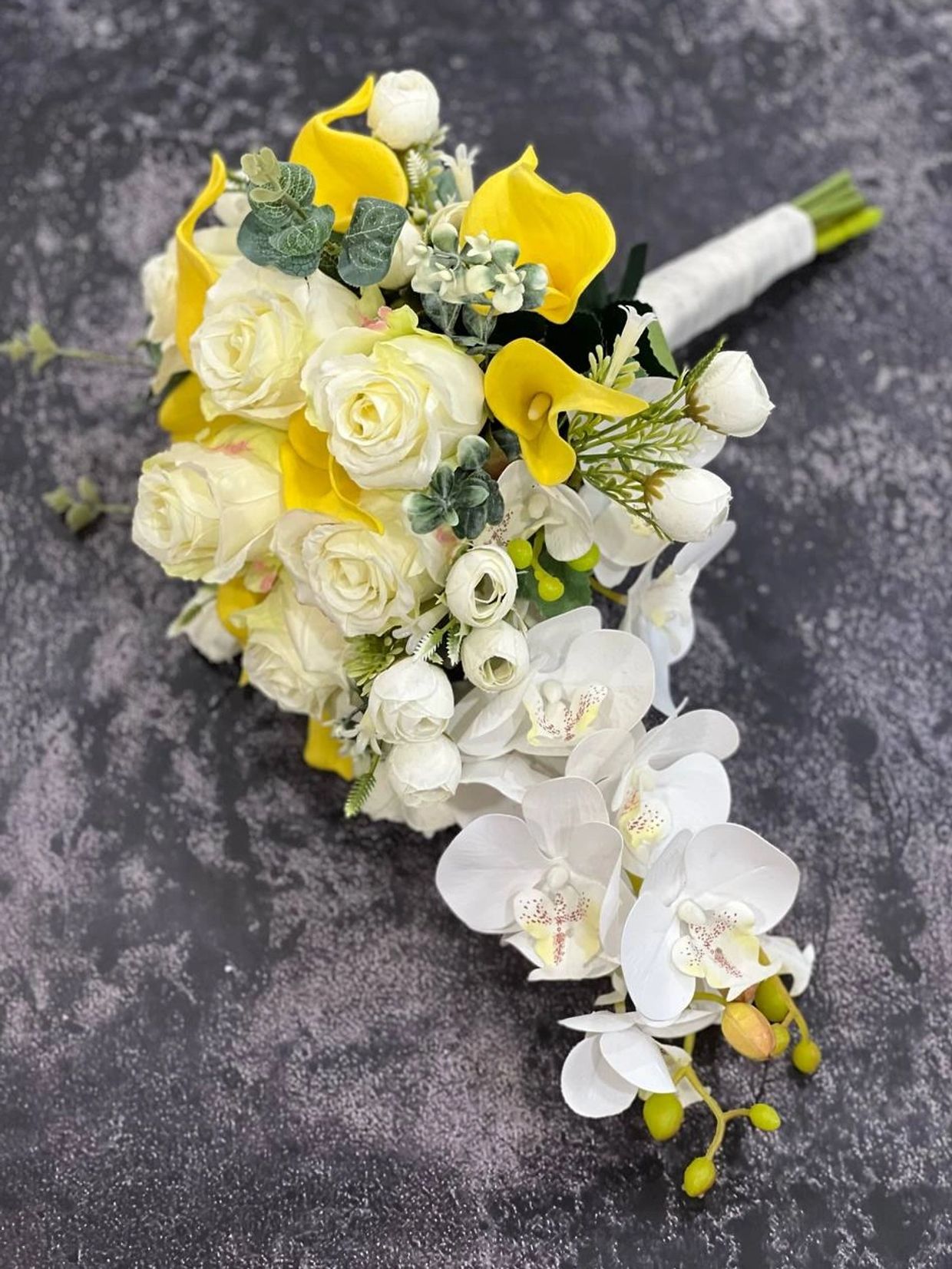 Elegant bouquet of white roses, yellow calla lilies, and white orchids on a dark textured surface.