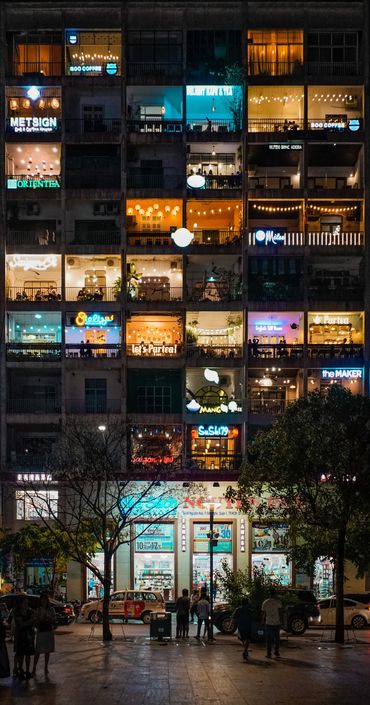A tall building with colorful lit windows at night and people walking outside.