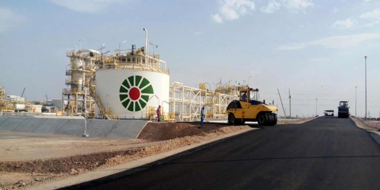 Heavy machinery paves a road near a large industrial tank with a green and red logo.