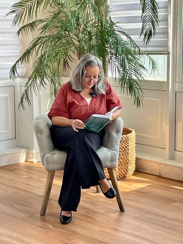 A woman reading a book on a chair near a large indoor plant.