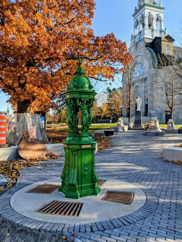 A bright green ornate water fountain in a park with autumn trees and a church in the background.