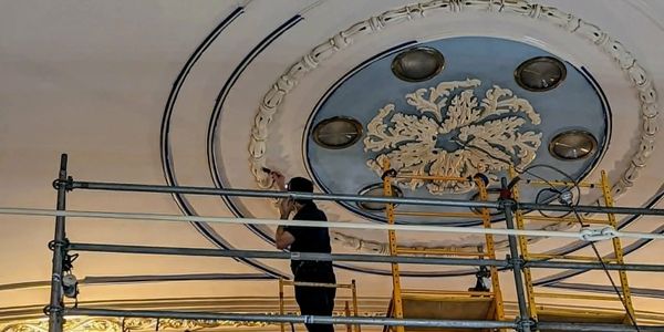 Worker on scaffolding near ornate ceiling medallion.