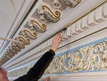 Person restoring intricate gold and white ornamental molding on a ceiling.