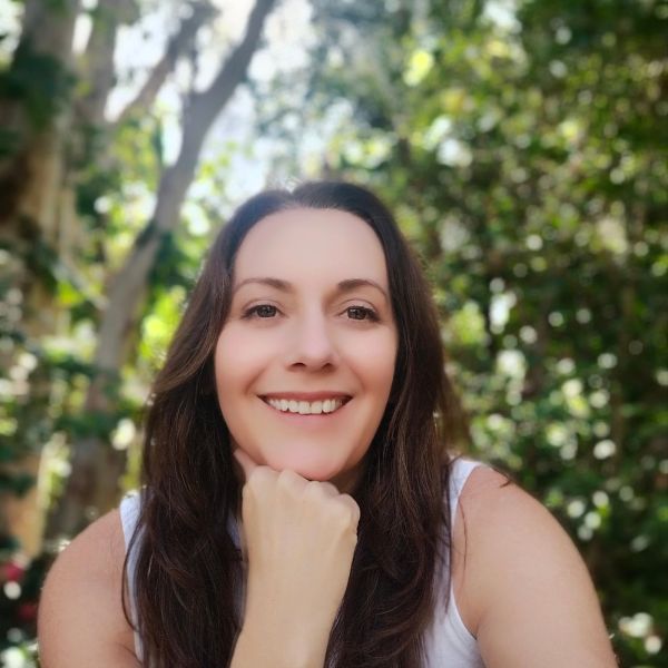 Smiling woman resting her chin on her hand outdoors with greenery in the background.