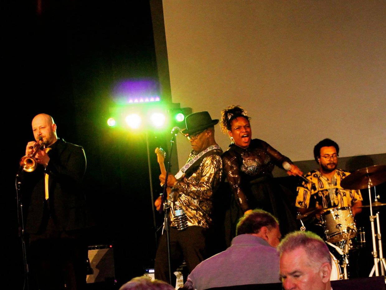 Band performing live with trumpet, guitar, vocals, and drums on stage.