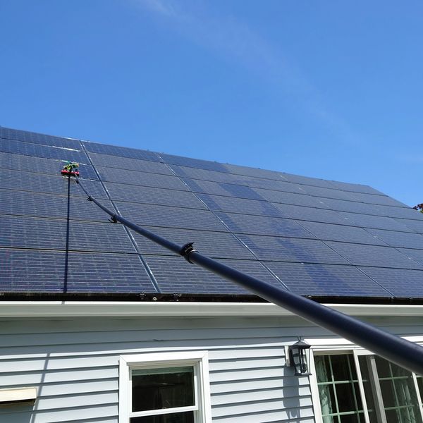 Cleaning solar panels on a house roof using an extended brush under clear blue sky.