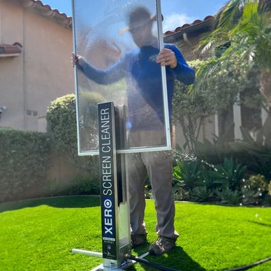 Person holding a window screen with Xero Screen Cleaner in a green garden.