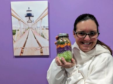 Woman smiling holding a colorful layered jar against a purple wall.