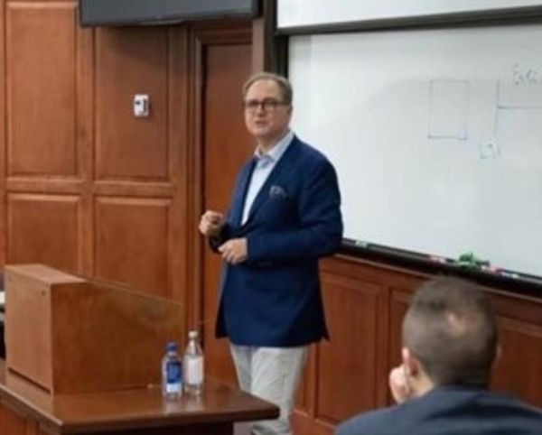 Man in blue blazer giving a lecture in a classroom with wooden panels.