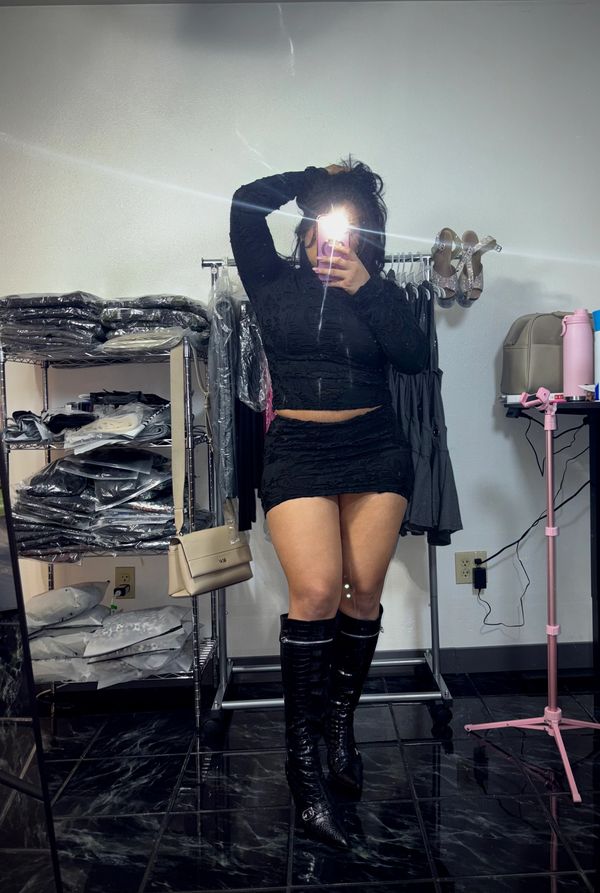 Person in black outfit and knee-high boots takes a mirror selfie in a room with clothing racks.