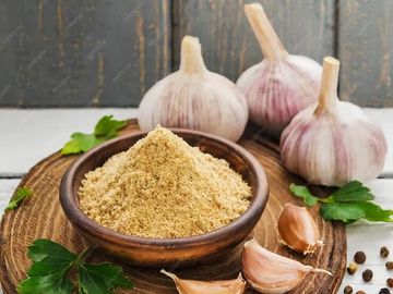 Ground garlic powder in a wooden bowl with garlic bulbs and parsley on a wooden board.