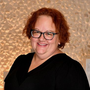 Smiling woman with glasses and curly red hair in black top.