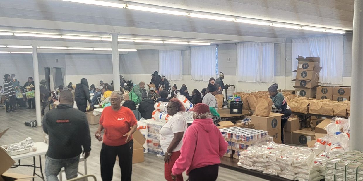 Community members gather indoors for a food distribution event with packed bags and supplies.
