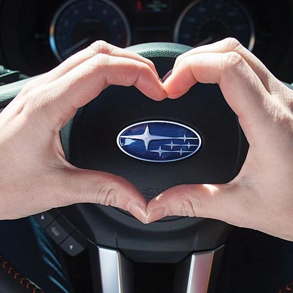 Hands forming a heart shape around a Subaru logo on a steering wheel.