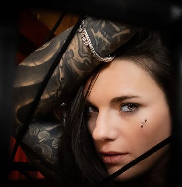 A woman with black hair, tattoos and facial piercings looks into the camera.