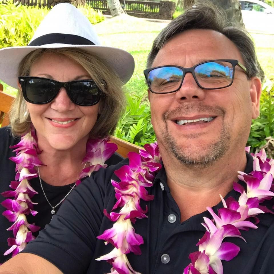 Smiling couple wearing flower leis and sunglasses, enjoying a sunny outdoor setting.